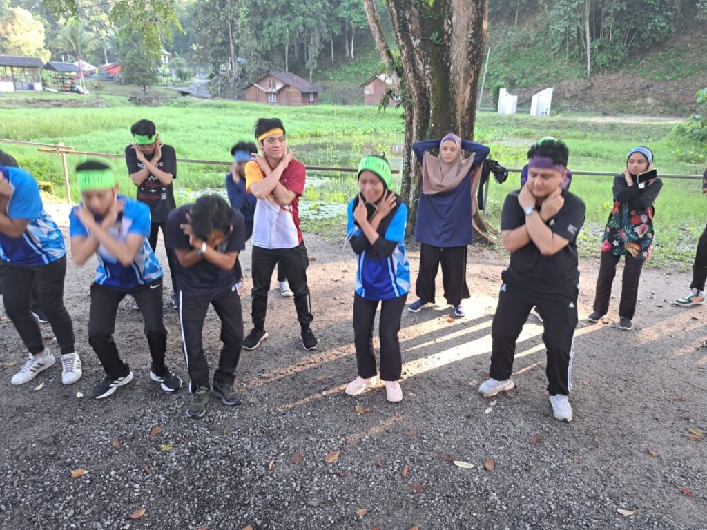 The image showcases JKM KSJ candidates engaging in a lighthearted punitive activity, promoting unity and memorization skills in a natural setting